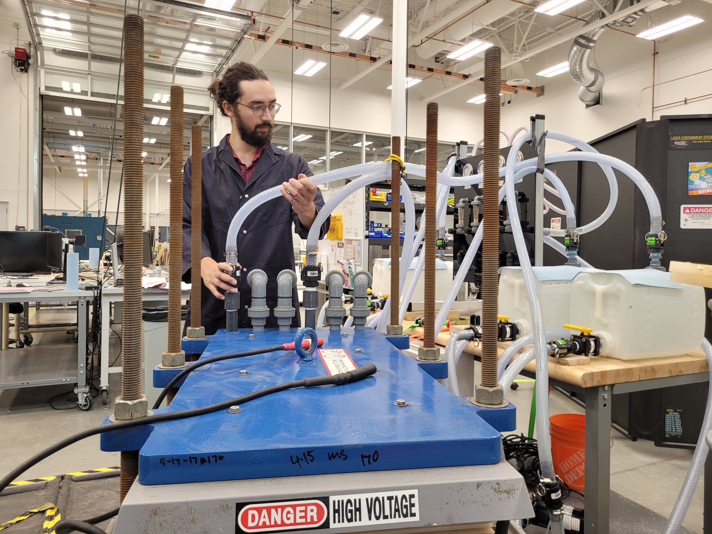A man in a lab coat and glasses stands in front a large flow battery with plastic tubes. 