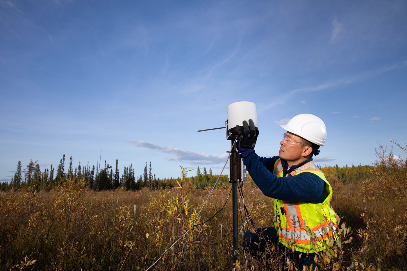 A man with a hard hard and hi-vis vest holds a clumb of peat and is standing in front of a vast peatland