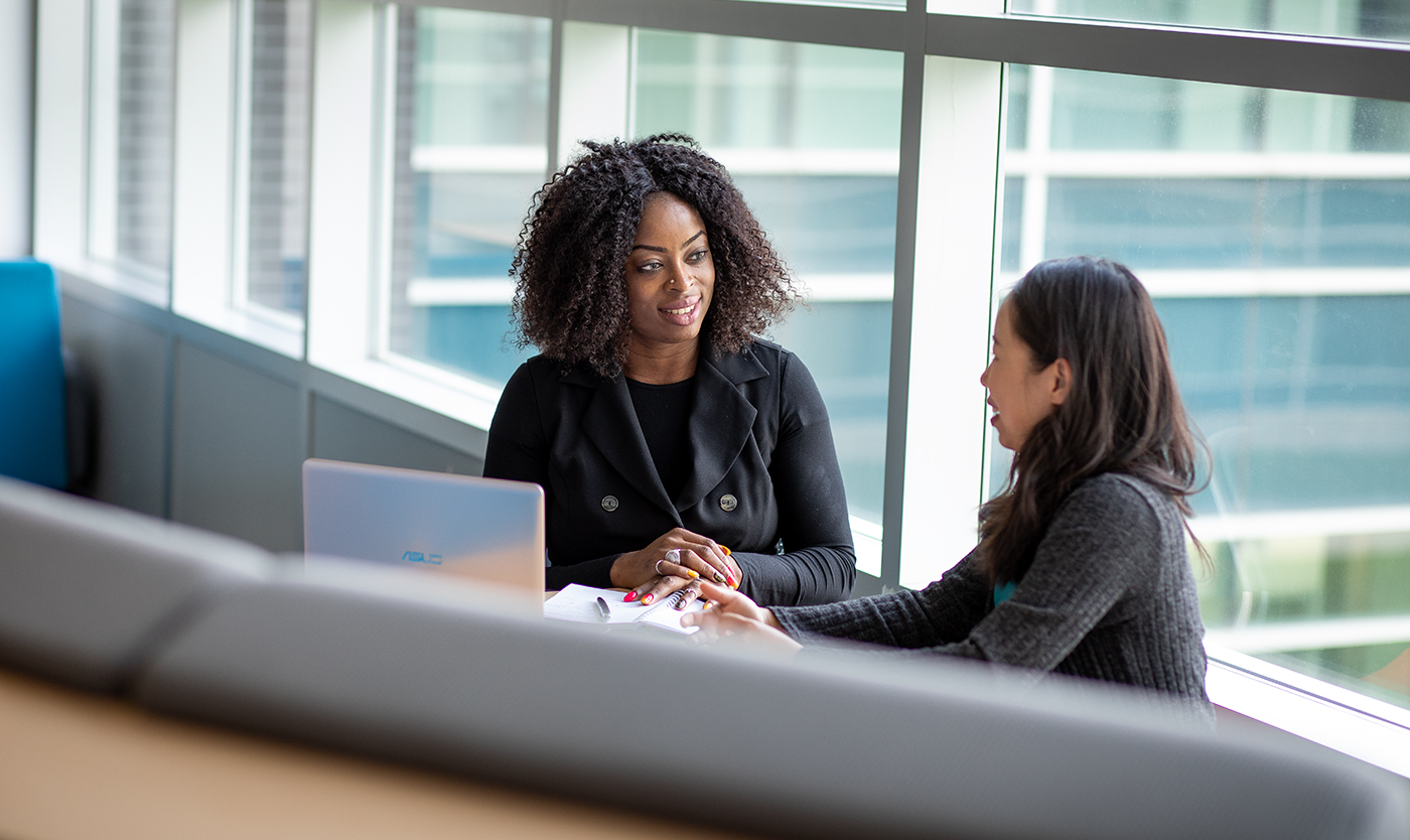 Woman with computer holding pen chatting with another woman at a table