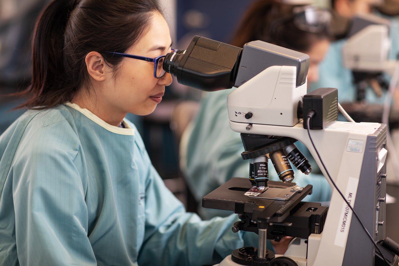 a woman wearing scrubs and eye protection and looking through a microscope
