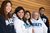 Five grads smile together in front of a wood panel wall all wearing white and blue NAIT sweatshirts