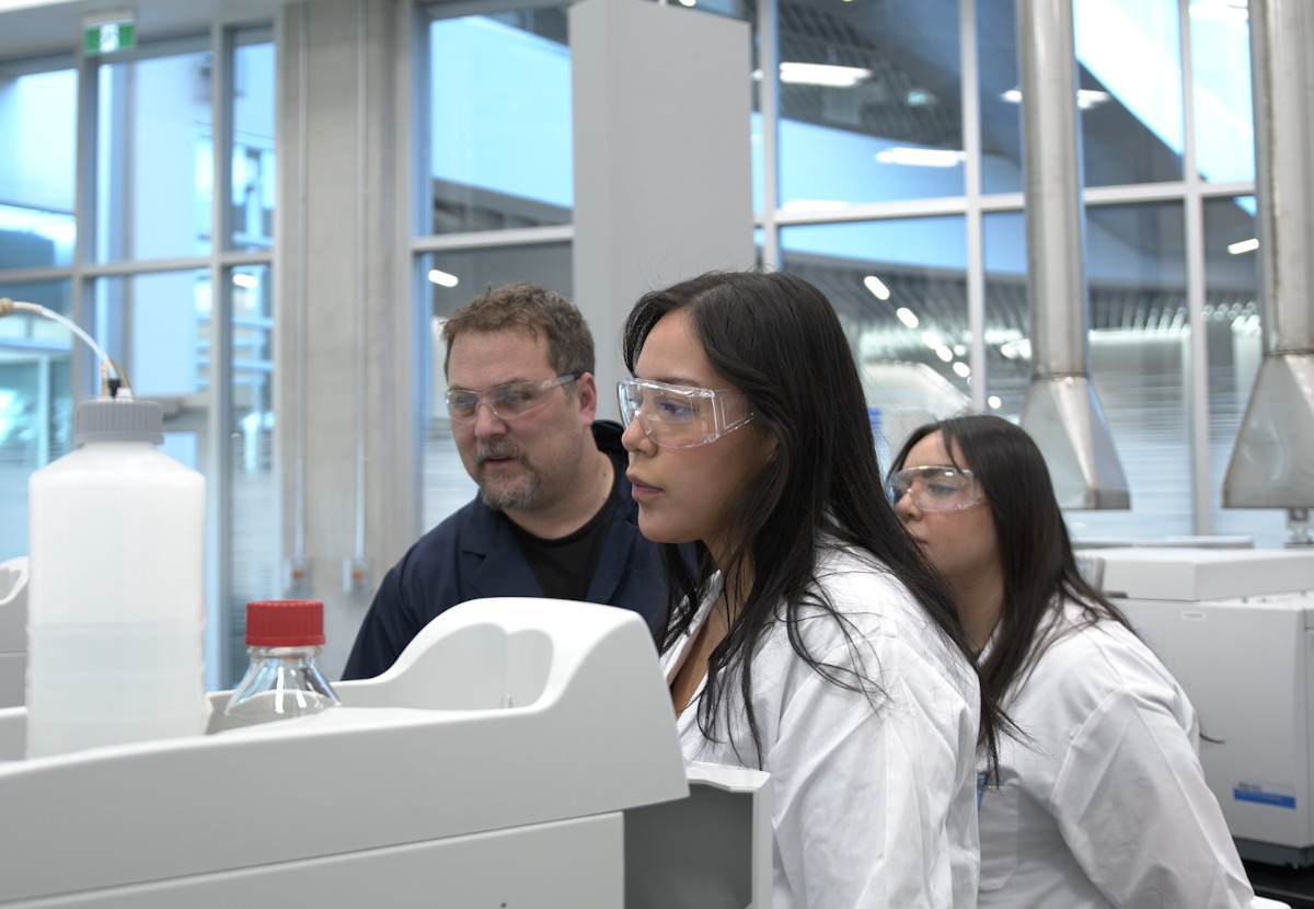 Two female students in lab coats and safety glasses look at the results of environmental testing while a professor watches