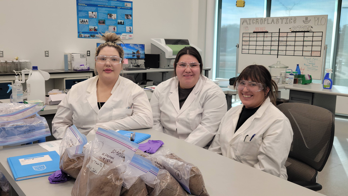 Three ladies from Peavine metis community smile. in lab coats and goggles and are in a clean technologies lab. 