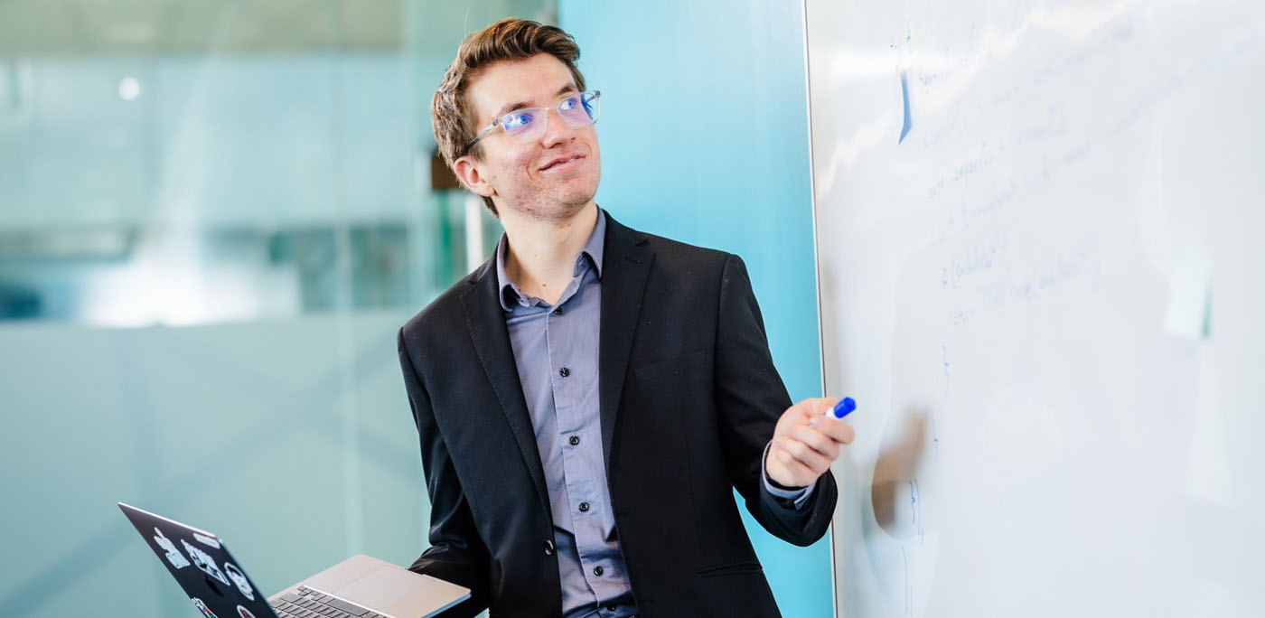 Man holding a laptop pointing to a whiteboard.