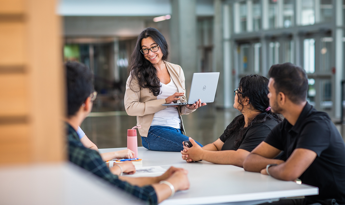 Woman in blazer holding laptop standing talking to group of people seated at a table